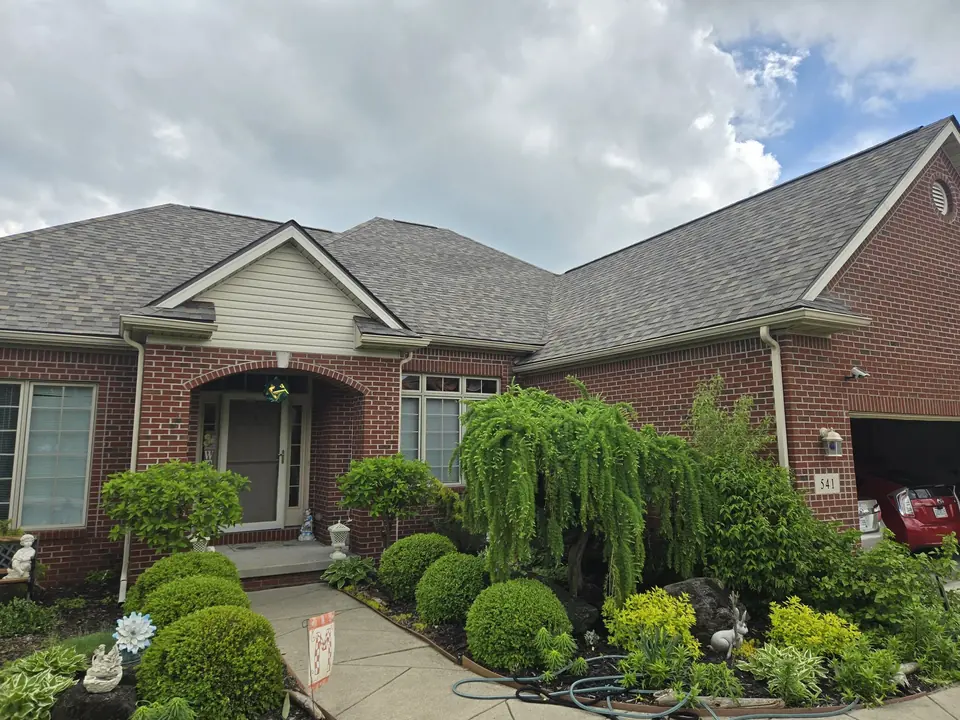 Brick home with a gray shingle roof above a landscaped front garden.