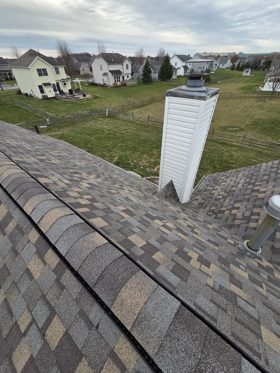 Ridge of a shingle roof with a siding-wrapped chimney, looking out over a subdivision.